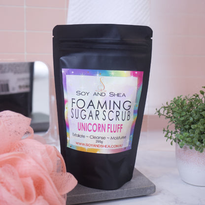 A black zip lock pouch stands upright on a stone tray in front of blurred bathroom background with peach tiles.   The pouch, which stands between a potted plant and a loofah, has a label that reads FOAMING SUGAR SCRUB UNICORN FLUFF and features a bright rainbow background