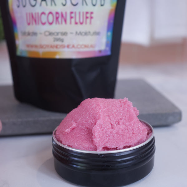 A zoomed in image of a black tin filled with pink sugar scrub sitting on a white bench with the blurred image of the packaging in the background.