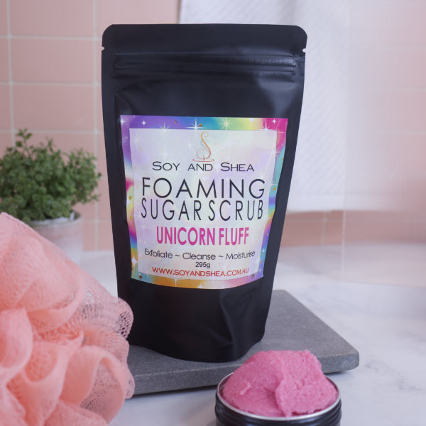 A black zip lock pouch stands up-right on a grey stone tray in front of pink tiled wall.  The Label reads  FOAMING SUGAR SCRUB UNICORN FLUFF against a bright rainbow background.    In front is a small black tin overflowing with a pink scrub .