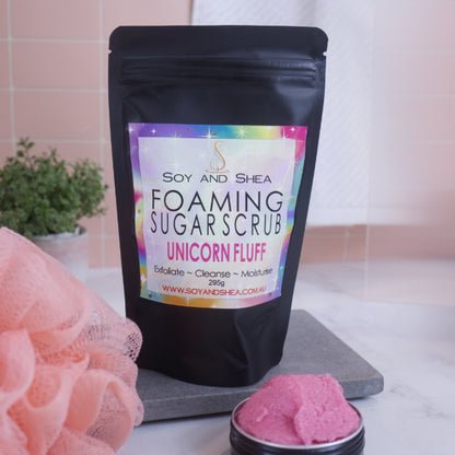 A black zip lock pouch stands up-right on a grey stone tray in front of pink tiled wall.  The Label reads  FOAMING SUGAR SCRUB UNICORN FLUFF against a bright rainbow background.    In front is a small black tin overflowing with a pink scrub .