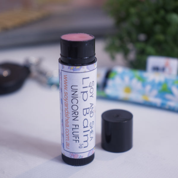 A black tube with colourful label reading LIP BALM UNICORN FLUFF stands upright on a white counter with a blurred background.  The black cap has been removed and sits to the side revealing a pink balm