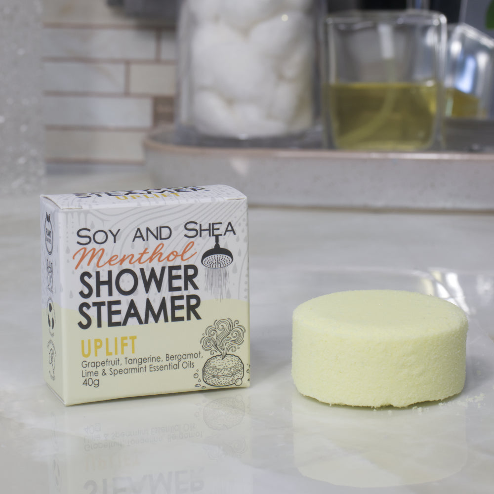 A boxed shower steamer and the steamer itself on a countertop in a bathroom setting. The box reads 'Soy and Shea Menthol Shower Steamer Uplift' and the round steamer is a pale yellow color.