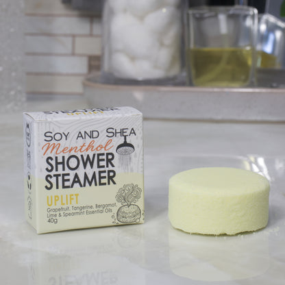 A boxed shower steamer and the steamer itself on a countertop in a bathroom setting. The box reads 'Soy and Shea Menthol Shower Steamer Uplift' and the round steamer is a pale yellow color.
