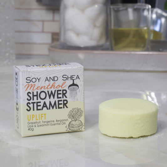 A boxed shower steamer and the steamer itself on a countertop in a bathroom setting. The box reads 'Soy and Shea Menthol Shower Steamer Uplift' and the round steamer is a pale yellow color.