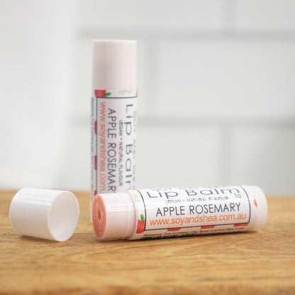 Two white tubes of lip balm are displayed on a wooden surface with blurred tiled background.  One tube stands on its end while the other lays flat in front with the cap removed revealing a light red balm.  The label is with a random pattern of red apples