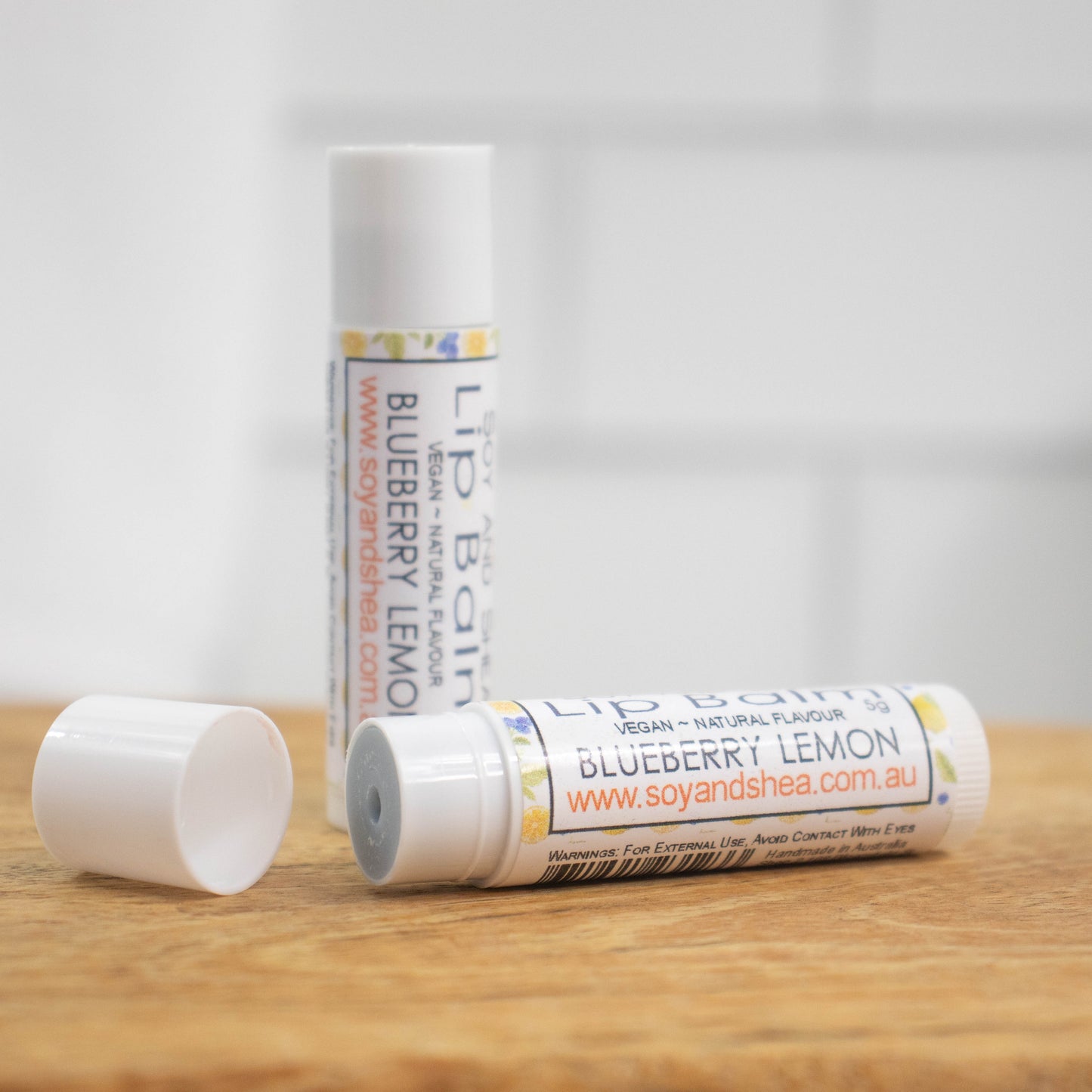 Two white tubes of lip balm are displayed on a wooden surface with blurred tiled background.  One tube stands on its end while the other lays flat in front with the cap removed revealing a light blue balm.  The label is white with a random pattern of lemon and blueberry illustrations