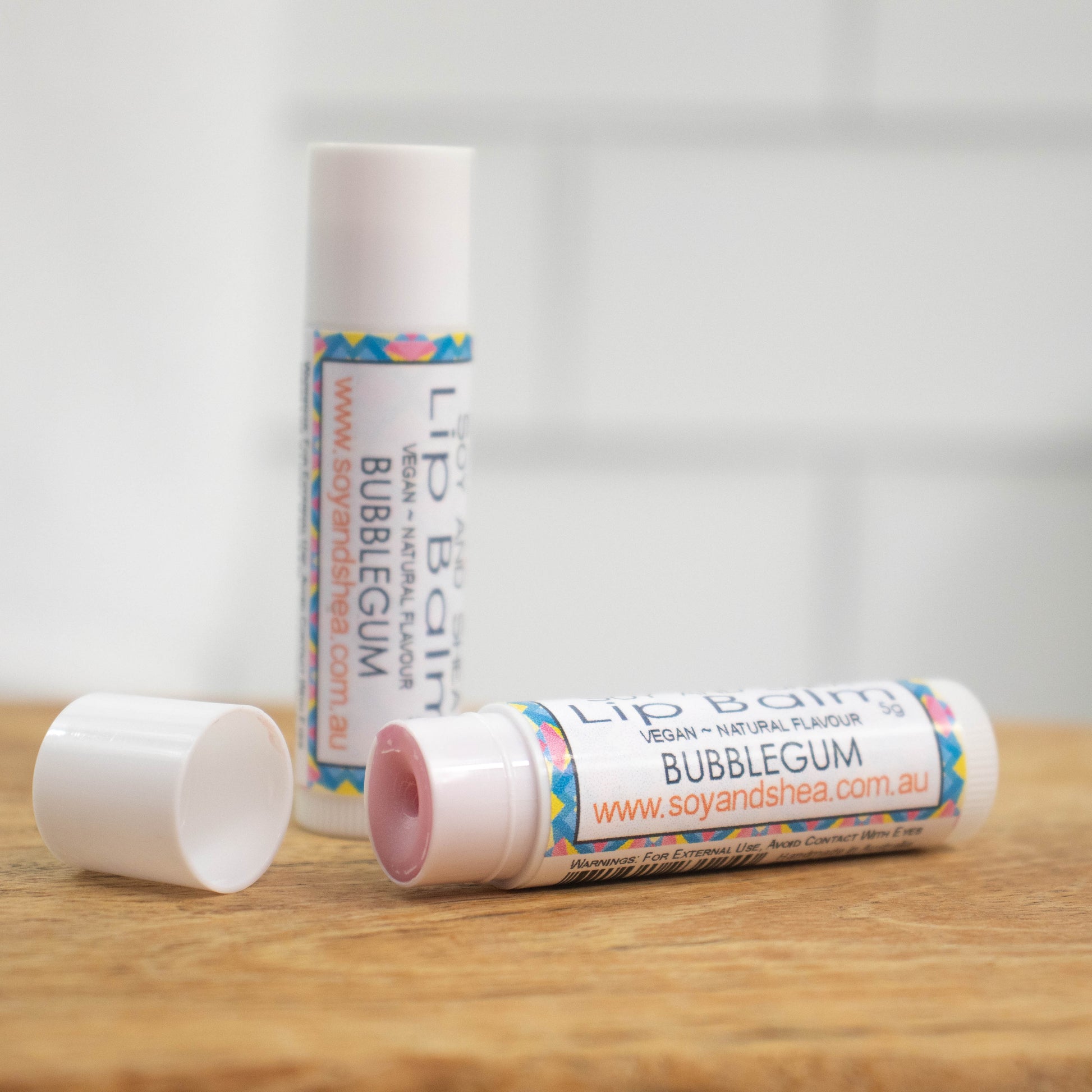 Two white tubes of lip balm are displayed on a wooden surface with blurred tiled background.  One tube stands on its end while the other lays flat in front with the cap removed revealing a pale pink balm.  The label has an abstract design in pink, yellow and blue