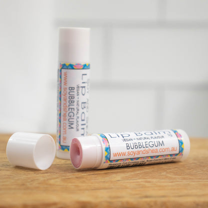 Two white tubes of lip balm are displayed on a wooden surface with blurred tiled background.  One tube stands on its end while the other lays flat in front with the cap removed revealing a pale pink balm.  The label has an abstract design in pink, yellow and blue