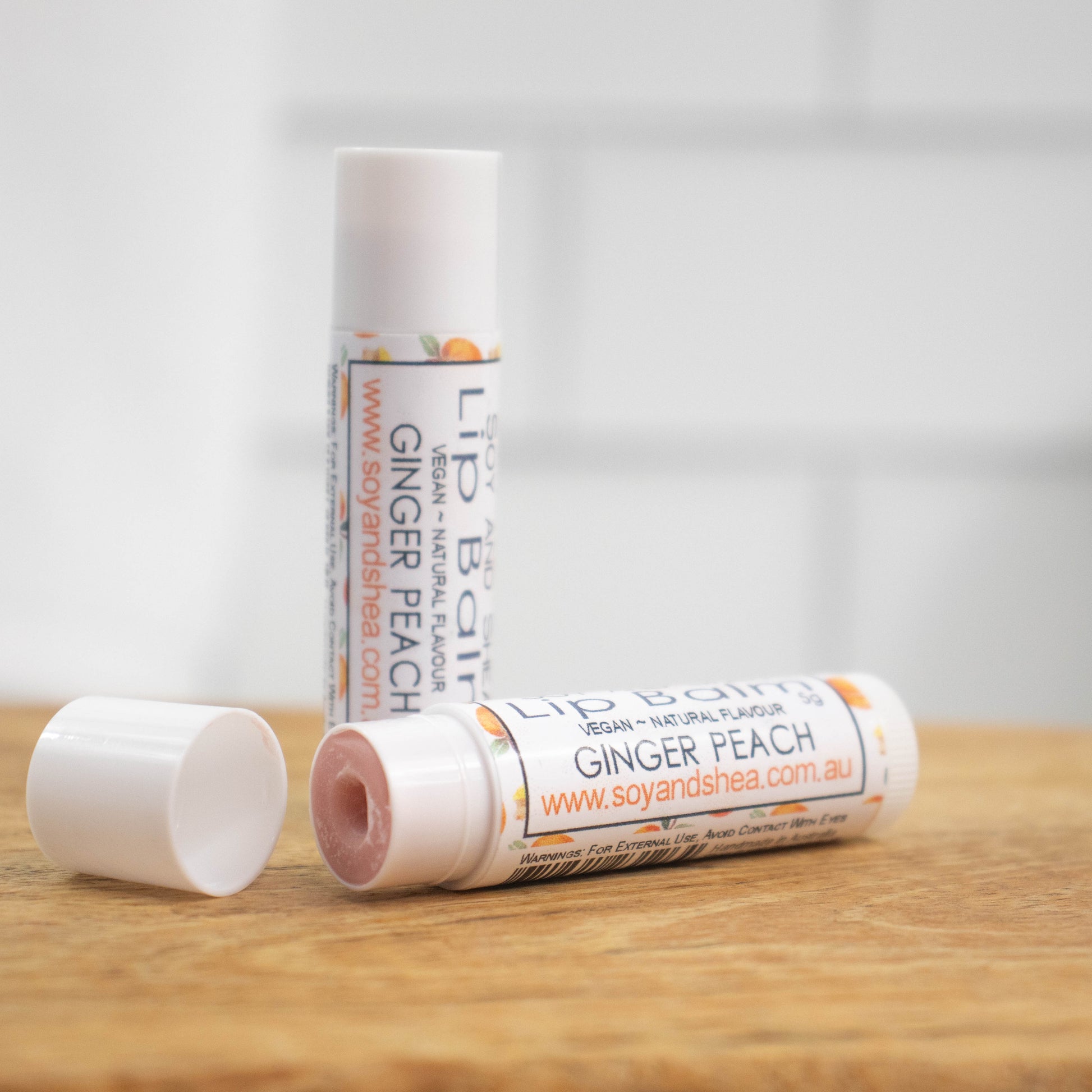 Two white tubes of lip balm are displayed on a wooden surface with blurred tiled background.  One tube stands on its end while the other lays flat in front with the cap removed revealing a light brown balm.  The label is white with peach and ginger pieces randomly scattered