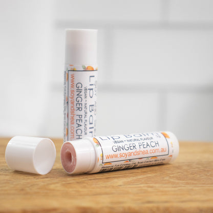Two white tubes of lip balm are displayed on a wooden surface with blurred tiled background.  One tube stands on its end while the other lays flat in front with the cap removed revealing a light brown balm.  The label is white with peach and ginger pieces randomly scattered