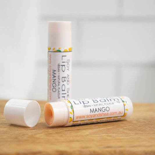 Two white tubes of lip balm are displayed on a wooden surface with blurred tiled background.  One tube stands on its end while the other lays flat in front with the cap removed revealing an orange balm.  The label is white with small illustration of whole mangos