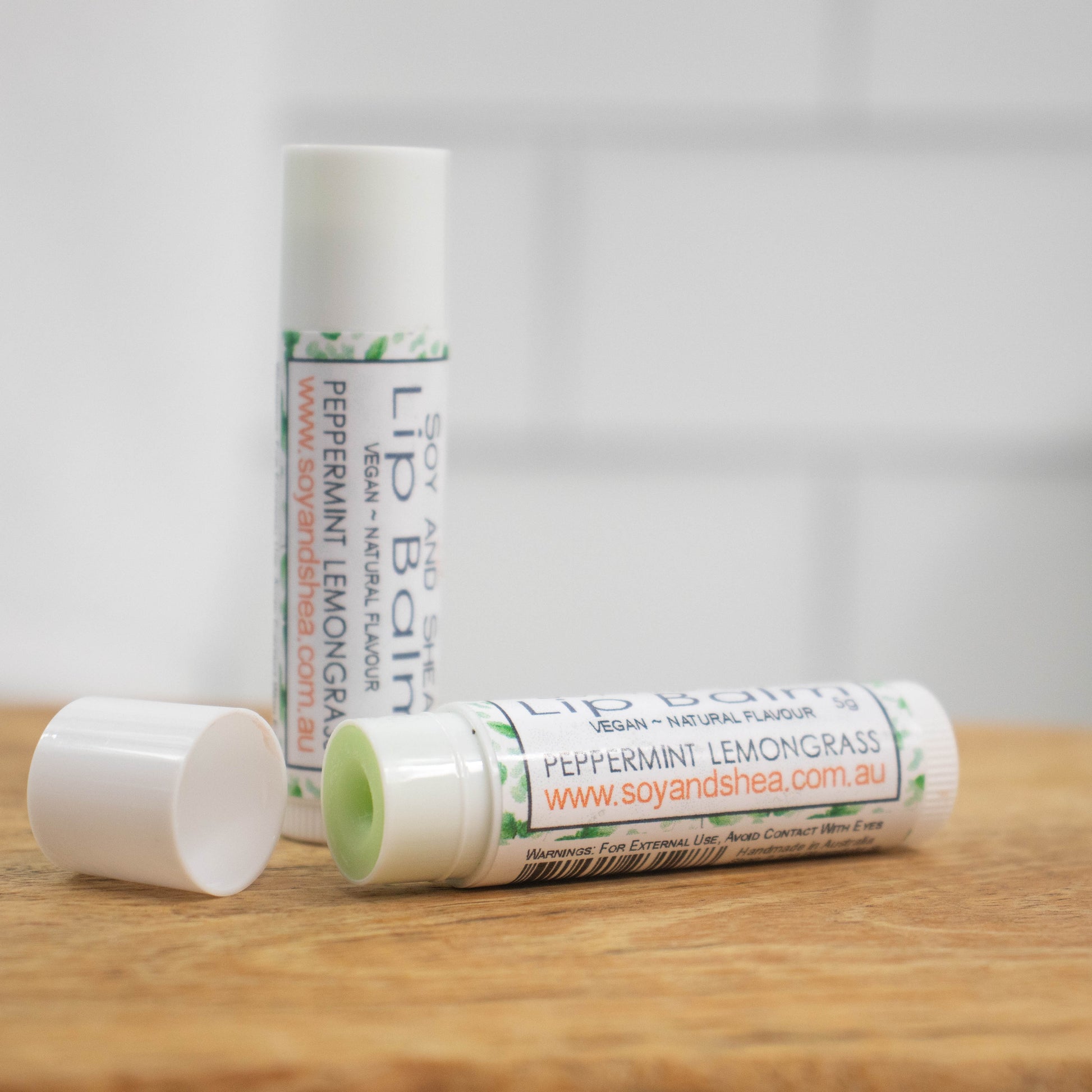 Two white tubes of lip balm are displayed on a wooden surface with blurred tiled background.  One tube stands on its end while the other lays flat in front with the cap removed revealing a light green balm.  The label is white with a green leaf pattern