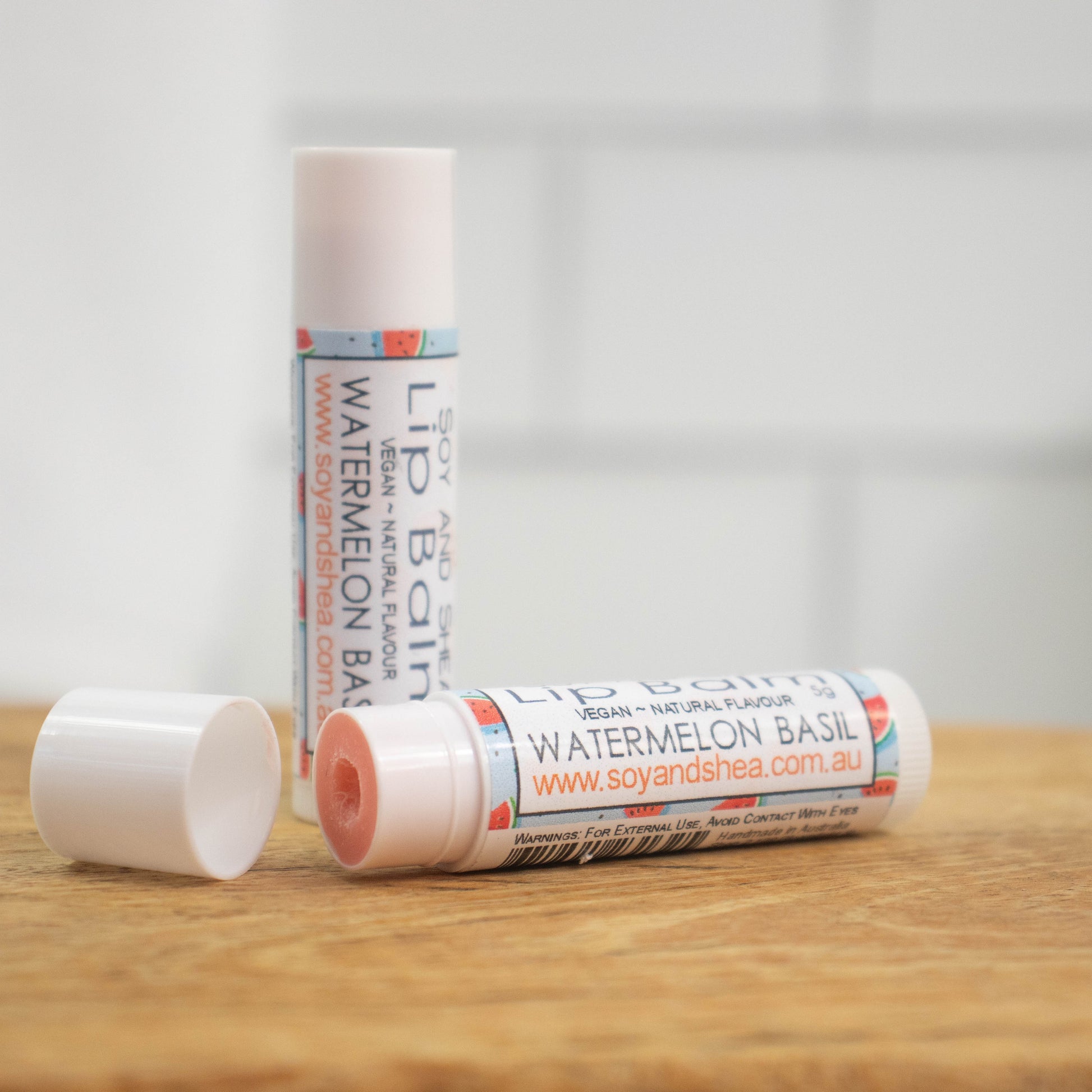 Two white tubes of lip balm are displayed on a wooden surface with blurred tiled background.  One tube stands on its end while the other lays flat in front with the cap removed revealing a light red balm.  The label has a blue background with random wedges of watermelon