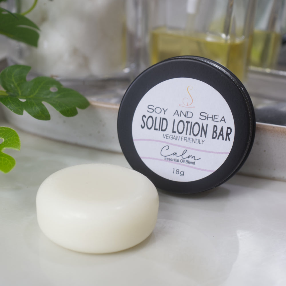 A small black tin with a label reading SOLID LOTION BAR stands on its side with the label facing forward.  In front of the tin is a solid white, cylindrical puck of lotion.  The background is blurred with greenery.