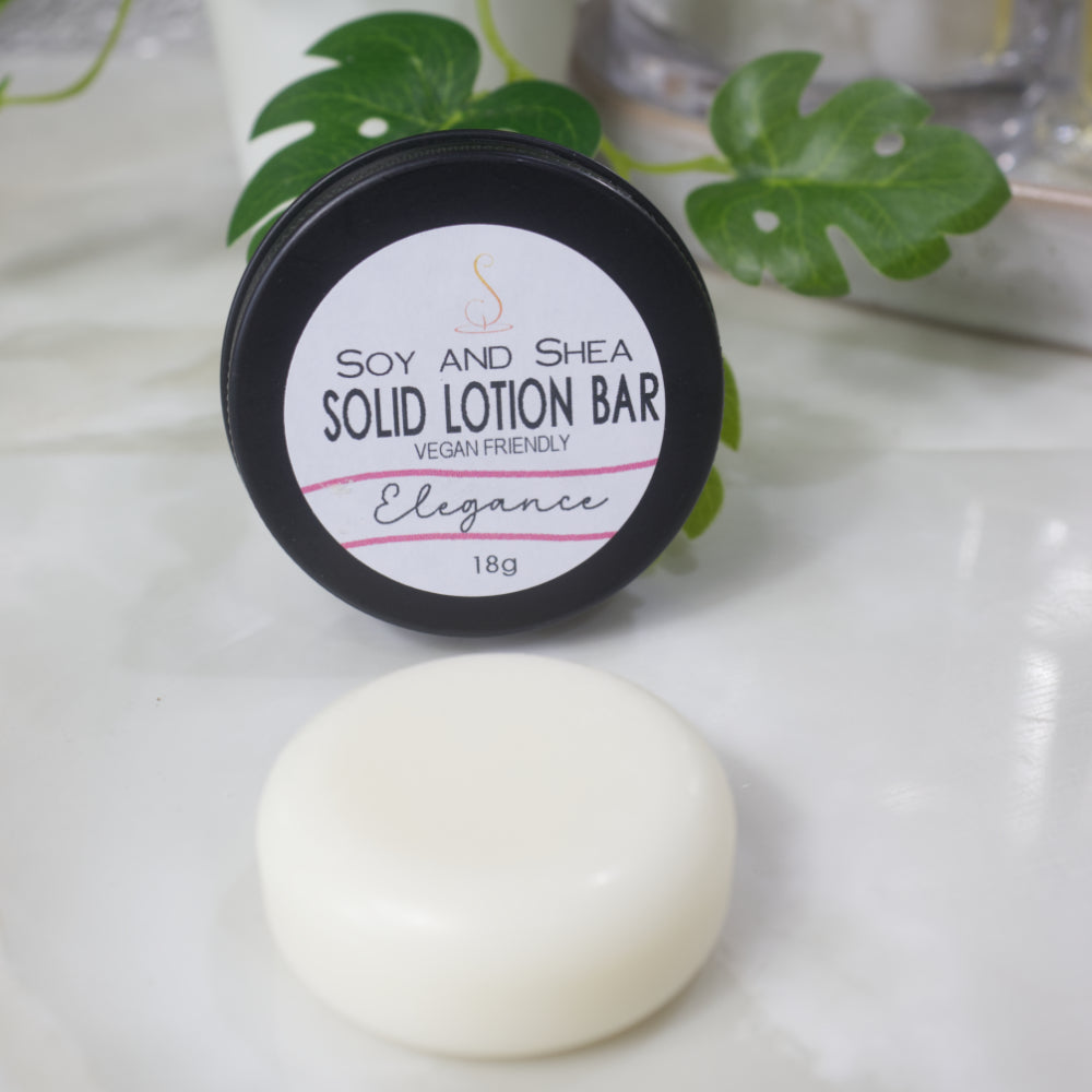 A small round black tin rests on its side with the label facing forward reading Solid Lotion Bar.  In front of the tin is a solid round puck of lotion.  In the background is a potted plant.