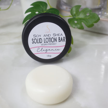 A small round black tin rests on its side with the label facing forward reading Solid Lotion Bar.  In front of the tin is a solid round puck of lotion.  In the background is a potted plant.