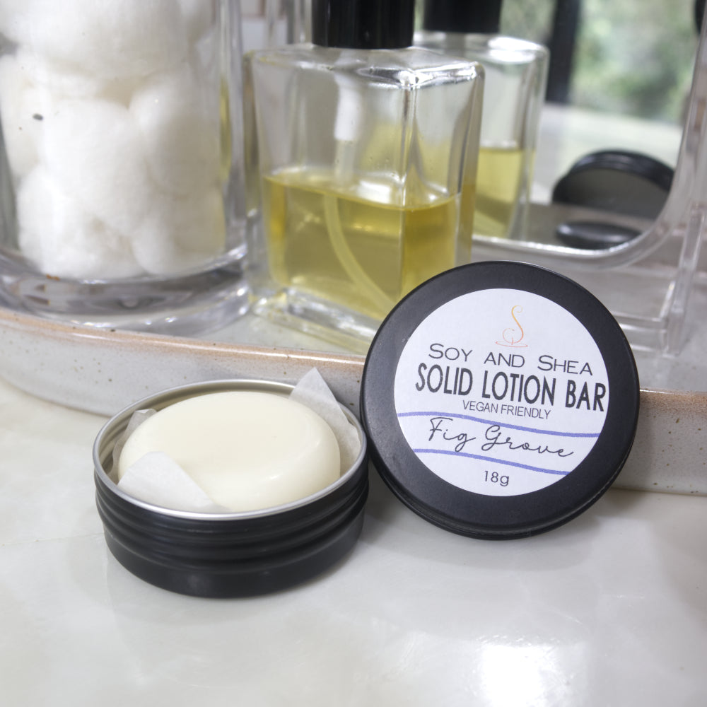 A small black tin sits ona marble bathroom counter surrounded by a stone tray with small mirror, Perfume bottle and jar of cotton balls.  The lid of the tin has been removed and rests to the side and features a lable reading SOLID LOTION BAR.  Inside the tin, is a white cylindrical puck