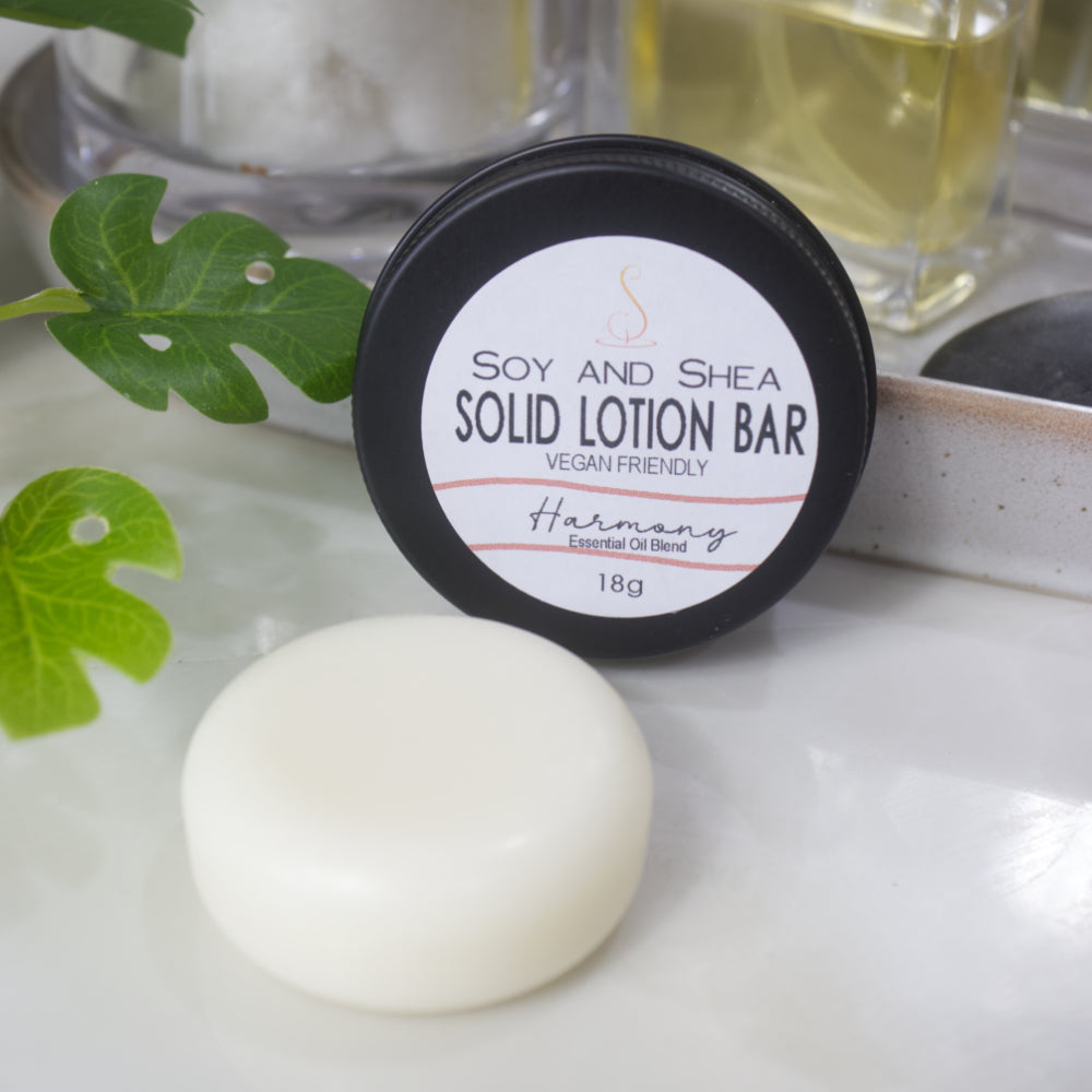 A small black tin with a label reading SOLID LOTION BAR stands on its side with the label facing forward.  In front of the tin is a solid white, cylindrical puck of lotion.  The background is blurred with greenery.