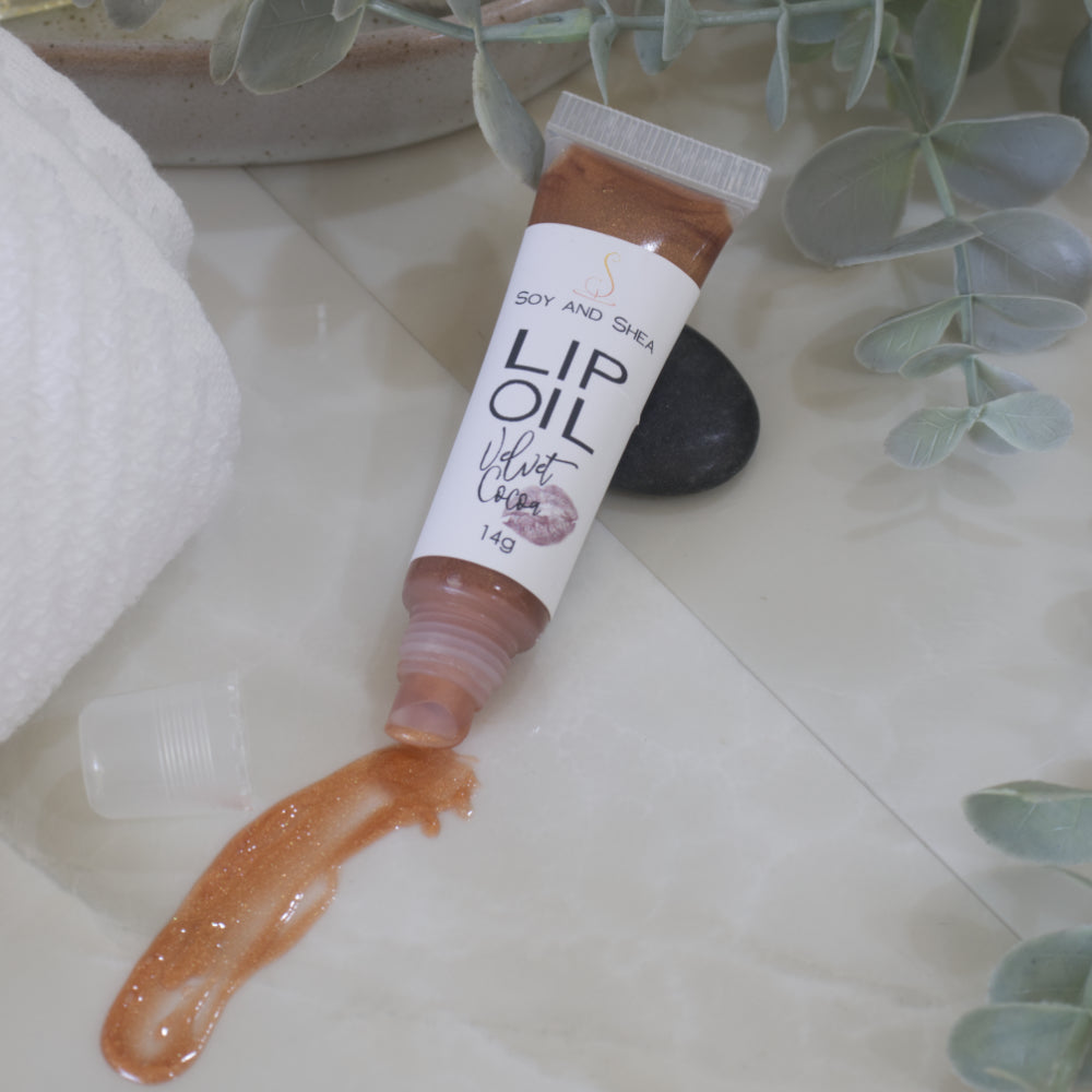 A tube of Lip Oil lies on a marble tile surface next to a white towel and some eucalyptus leaves.   The tube is clear with a white product label and filled with a chocolate brown Lip oil.  The cap is removed and a smear of Lip oil is on the bench showing the translucent effect it has