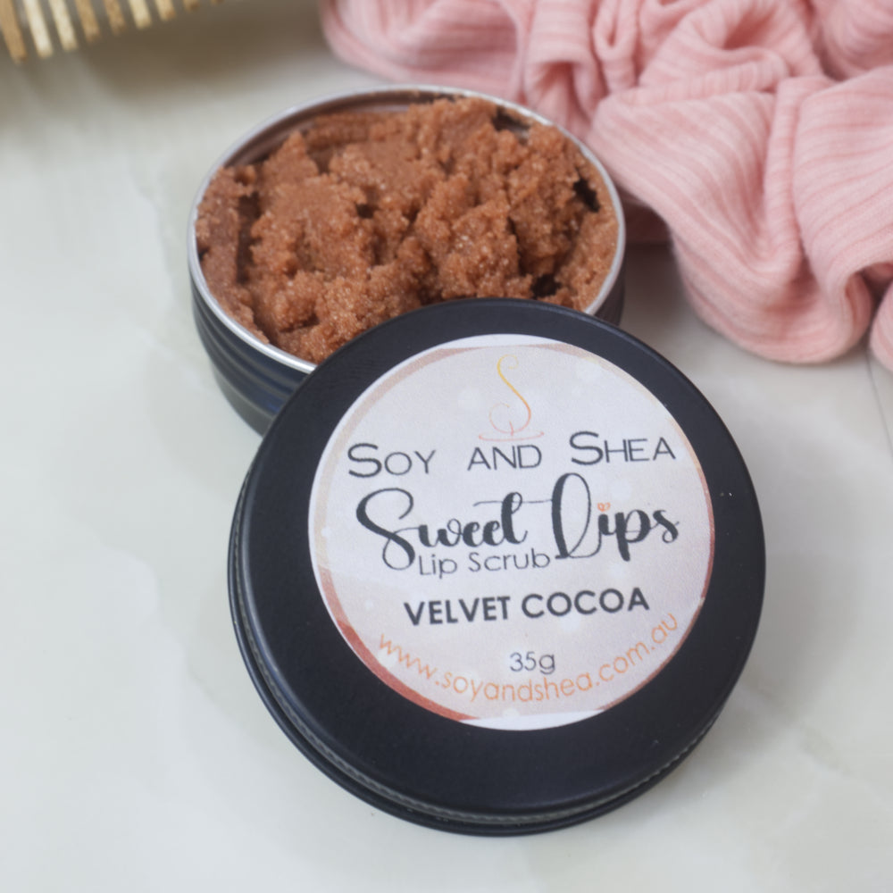 A black tin of Lip Scrub sits on a marble countertop next to a scrunchie and wooden comb.  The lid has been removed and rests on the side of the tin revealing a chocolate brown scrub