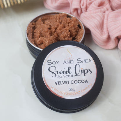 A black tin of Lip Scrub sits on a marble countertop next to a scrunchie and wooden comb.  The lid has been removed and rests on the side of the tin revealing a chocolate brown scrub