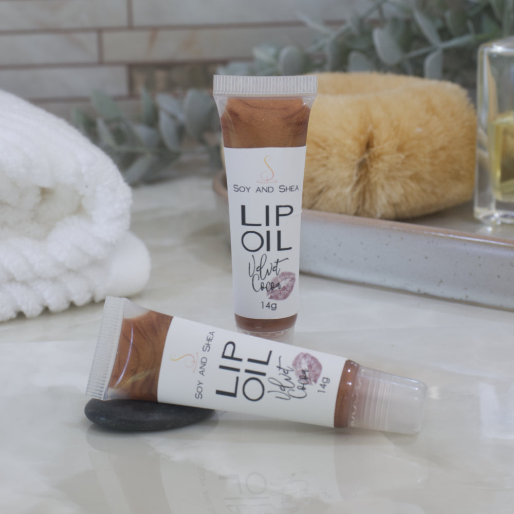 Two tubes of Lip Oil are presented on a bathroom countertop with a blurred background of towels, greenery and body brush.   One tube stands upright while the other lays flat in front of it.  The tubes are clear with a white product lable and are filled with a chocolate brown Lip oil.