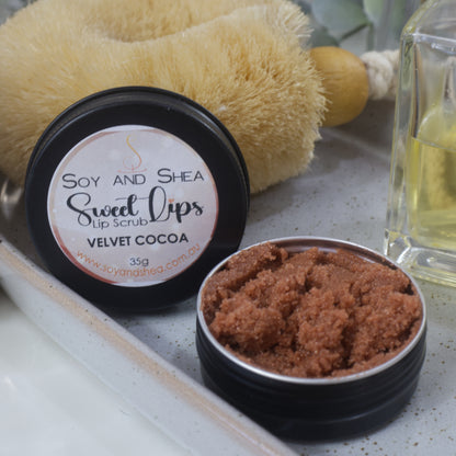 A round black tin sits on a stone tray along with a bottle of perfume and body brush.  The lid has been removed and is resting against the body brush revealing a chocolate brown sugar scrub 