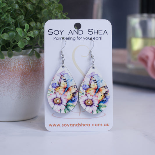 A pair of earrings on silver hooks hang on a white card branded with SOY AND SHEA leaning against a pot with a green leafy plant and a blurred background.  The earrings are teardrop shaped with a watercolour image of a large orange butterfly surrounded by a pink flower and leaves