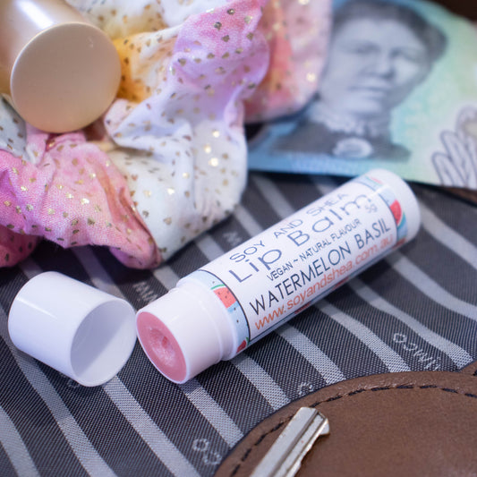 A white tube of lip balm lies on an open handbag with the cap off revealing a light red balm.  It is surrouded by a scrunchie and money.  The label is blue with illustrations of watermelon slices.