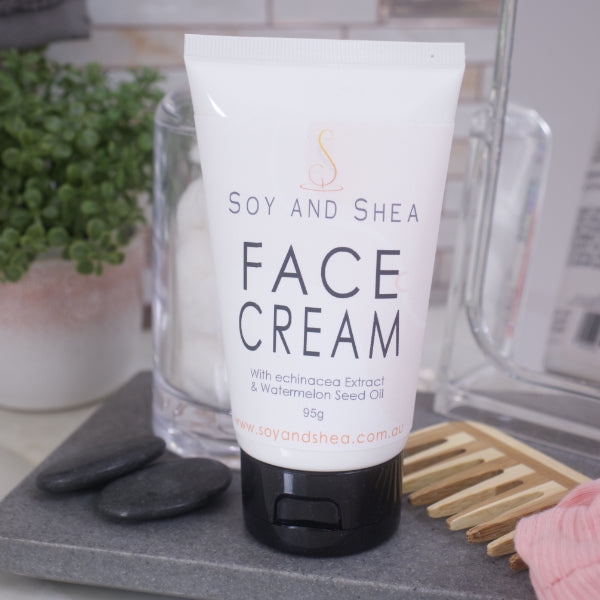 Tube of Soy and Shea Face Cream on a bathroom counter with a comb and stones.  The cream is in a white tube with black lid