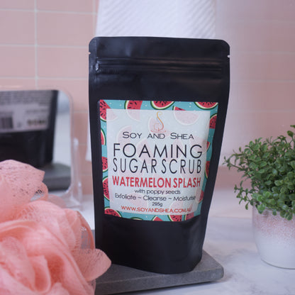 A black zip lock pouch stands upright on a stone tray in front of blurred bathroom background with peach tiles.   The pouch, which stands between a potted plant and a loofah, has a label that reads FOAMING SUGAR SCRUB WATERMELON SPLASH and features drawings of watermelon slices against a teal background