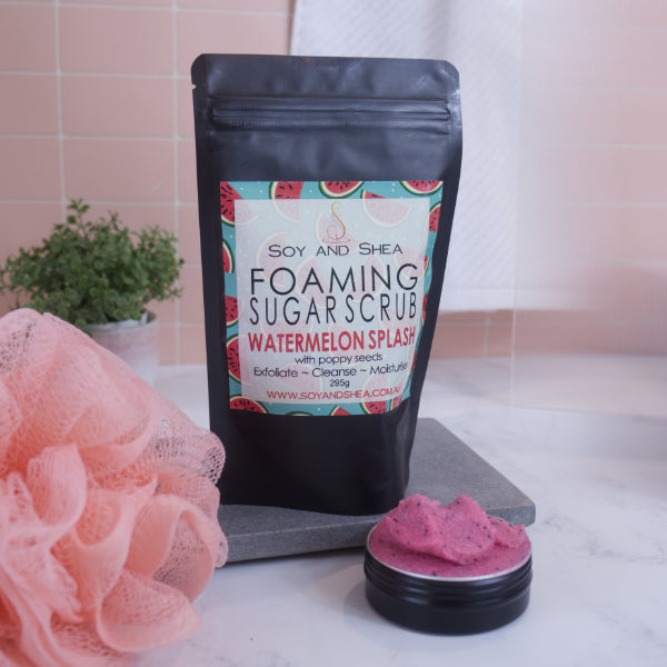 A black zip lock pouch stands up-right on a grey stone tray in front of pink tiled wall.  The Label reads  FOAMING SUGAR SCRUB WATERMELON SPLASH and has drawing of watermelon slices against a teal background.    In front is a small black tin overflowing with a pink scrub with black flecks of poppy seeds.