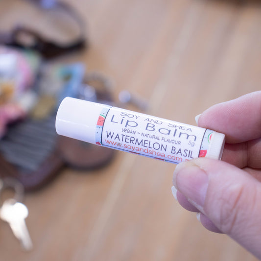A hand holds a white tube labelled lip balm over a blurred image of an open handbag with contents spilling onto the table.  The lip balms features a blue label with watermelon slices