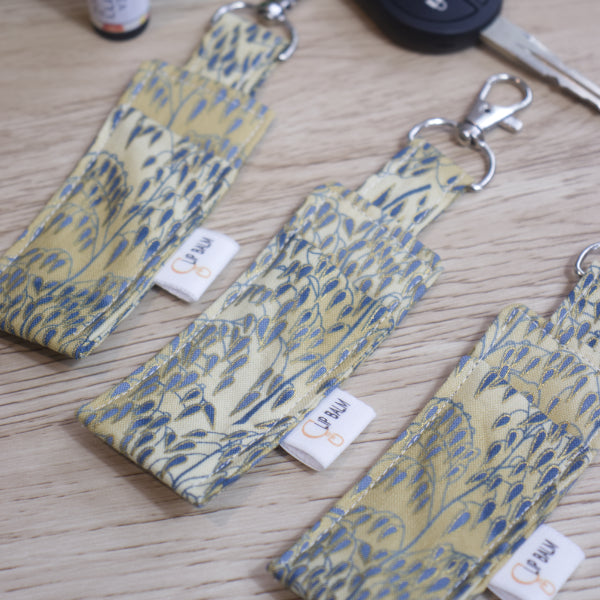 A selection of three lip balms showing variations in fabric design, lie on a wooden side  with a car key near-by.  The Clip balms have a mottled cream background that is covered with branches of weeping willow with blue leaves outlined in gold