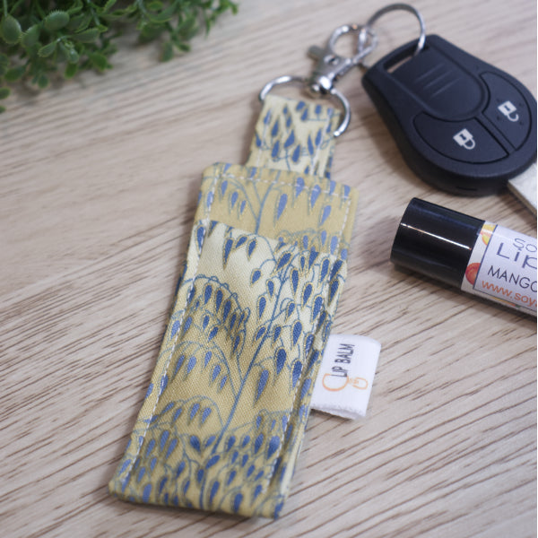 A Lip Balm holder lies on an wooden surface with lobster claw clasp attached to a car key.  A lip balm and greenery can be seen in the image.  The Clip Balm has an illustration of weeping willow branches with blue leaves outlined in gold agains a mottled cream background