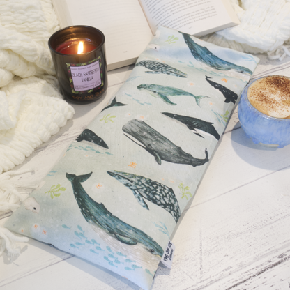 A long rectangular heat pack depicting a variery of watercolour whale species on a pale blue background rest  on a white blanket and open book, displayed next to a candle and a cup of coffee.
