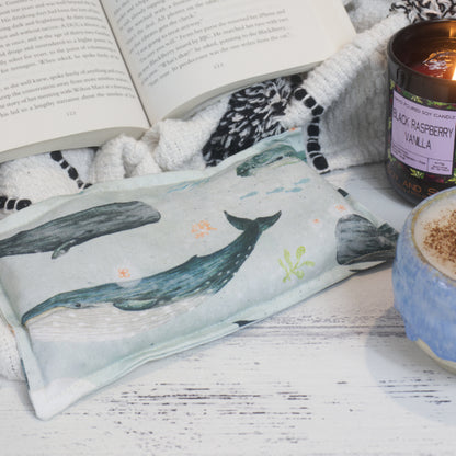 A Whales Mini Heat Pack rests on a table top next to an open book, burning candle and filled coffee cup creating a cosy scene.   The Mini Heat Pack is rectangular and features watercolour drawings of various what species on a pale blue background