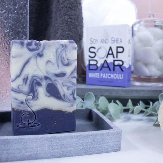 A tall rectangular bar of soap sits on a grey soap dish. The base of the soap is a rich purple with a thin gold line on top. This is followed by a white layer with rich purple swirls.  Behind the soap is a shower screen and towel along with a box the soap was presented in. The box has a white top with purple bubble border.