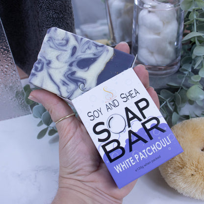 A hand is holding onto a box that has a bar of soap coming out the top. The lower half of the box has a purple bubble shaped border, and the top is white with grey bubbles. The box shows it is soap and the scent. The soap is rectangular in shape with a layered rich purple bottom, with fold line and the top is white with purple swirls.