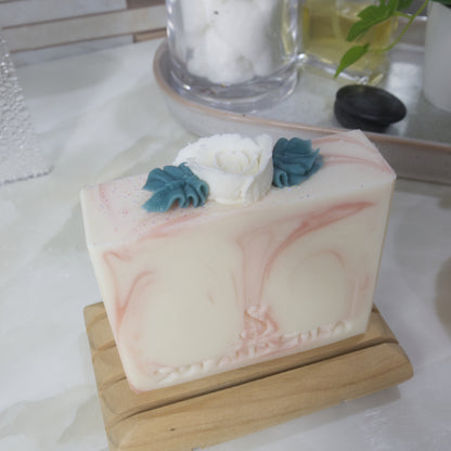 A white bar of soap with pink swirls is standing on a white marble bench with a blurred background.  The images focuses on the soap top which features a white rose with green leaves piped with soap.
