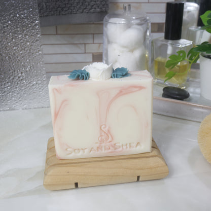 A bar of soap is standing on a wooden soap rack on a bathroom countertop.  The soap is white with delicate light pink swirls.  The top of the soap features a white soap rose in the center with dark green leaves coming of the side of the rose