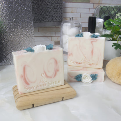 Three soaps are displayed on a bathroom countertop.  To the right, one soap lays flat showing the details of the soap top while another stands upon it.  The left front, is a third soap on a wooden soap rack.  The soaps have a main colour of white with varying light pink swirls.  Each soap features a white rose with green leaves on the top center.  These are made with soap