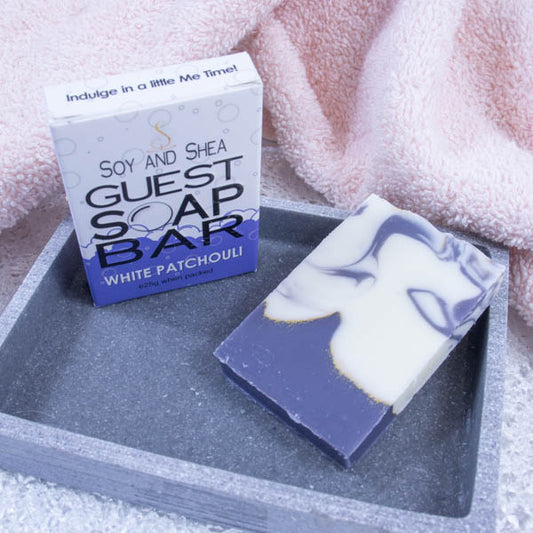 A grey stone soap dish sits next to a pink towel.  On the dish stands a white box with a border of purple bubble foam across the bottom and text GUEST SOAP BAR.   Next to the box lies a piece of soap that has a dark purple wavy base layer taking up about a quarter of the soap.  It is topped with a white soap with the same purple colour swirled throughout.