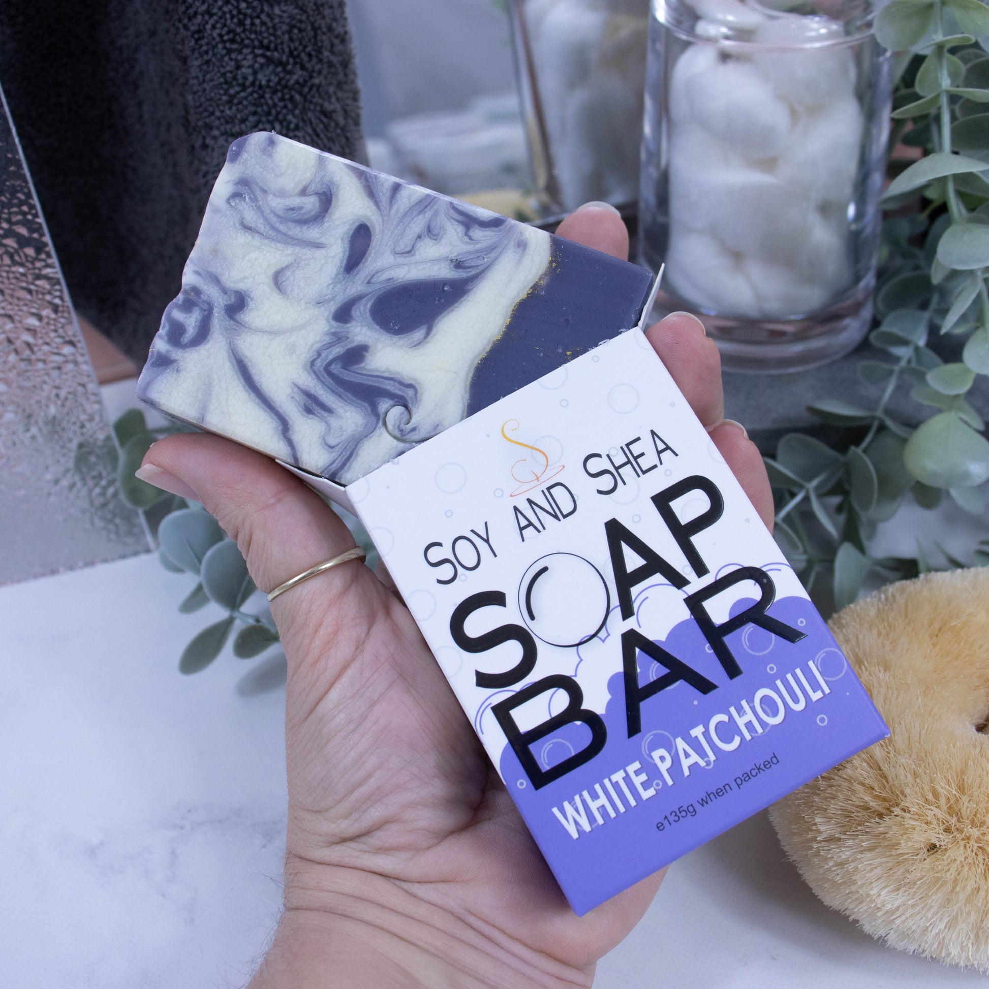 A hand is holding onto a box that has a bar of soap coming out the top. The lower half of the box has a purple bubble shaped border, and the top is white with grey bubbles. The box shows it is soap and the scent. The soap is rectangular in shape with a layered rich purple bottom, with gold line and the top is white with purple swirls.  In the background is a blurred bathroom counter top.