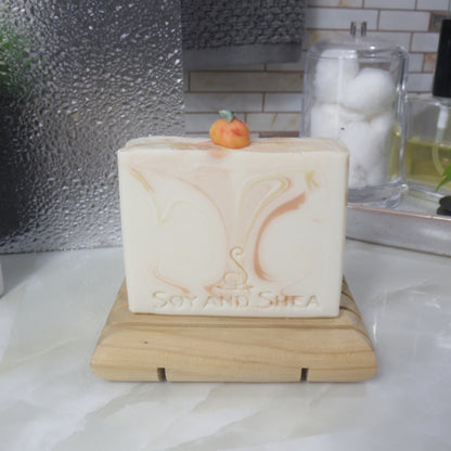 A soap stands on a wooden soap rack on a bathroom countertop.   The soap is a cream colour with wisps of orange and yellow throughout.  One top of the soap is a small replica of peach made with soap.