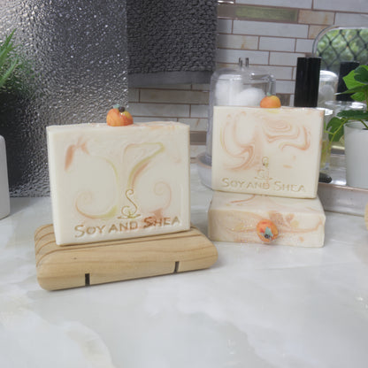 Three bars of soap are displayed on a bathroom countertop surrounded by plants, perfume and jar of cotton balls.   One soap stands the left on a wooden soap dish, while to the right, another soap lays flay on the surface with another standing ontop of it.   All soaps are a cream colour with varying wisps of orange and yellow swirls.  Each soap features a small peach decoration made of soap