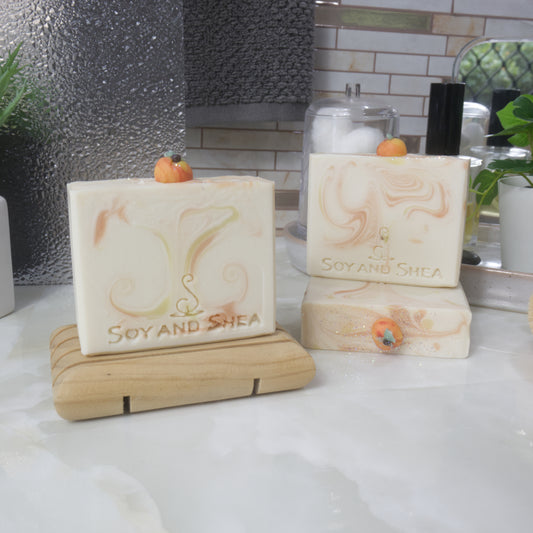 Three bars of soap are displayed on a bathroom countertop surrounded by plants, perfume and jar of cotton balls.   One soap stands the left on a wooden soap dish, while to the right, another soap lays flay on the surface with another standing ontop of it.   All soaps are a cream colour with varying wisps of orange and yellow swirls.  Each soap features a small peach decoration made of soap