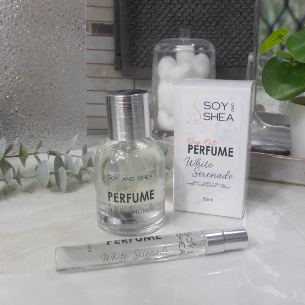 A selection of perfume bottles are presented on a bathroom countertop.  To the front is a clear, narrow, tall bottle labeled as 10ml with a silver cap and behind is a clear round, short bottle with silver lid labelled as 50ml.  Behind sits a white box with label identifying as Dry Oil Perfume White Serenade.