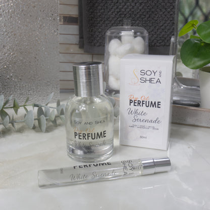 A selection of perfume bottles are presented on a bathroom countertop.  To the front is a clear, narrow, tall bottle labeled as 10ml with a silver cap and behind is a clear round, short bottle with silver lid labelled as 50ml.  Behind sits a white box with label identifying as Dry Oil Perfume White Serenade.