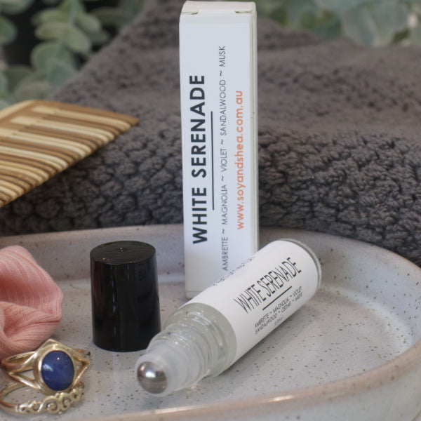 A small narrow clear bottle with the black lid removed and standing to the right revealing a metal roller mechanism rests on a stone tray with rings. Beside the bottle is a white box standing up right with WHITE SERENADE on the box and bottles label. A grey towel, greenery and wooden comb can be seen in the background.